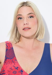 Le maillot de bain présente un motif floral en rouge et bleu, avec un décolleté en V profond. Le matériau est lisse et extensible.