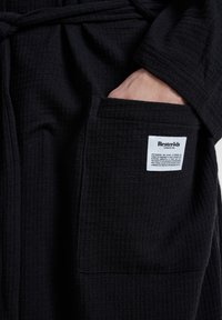 Bata negra con textura de waffle que muestra una mano en un bolsillo lateral. Presenta bordes cosidos y una etiqueta con los detalles del producto en el bolsillo.