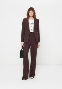 Femme en costume tailleur marron foncé et t-shirt imprimé blanc tenant un sac à main noir contre un mur blanc uni.