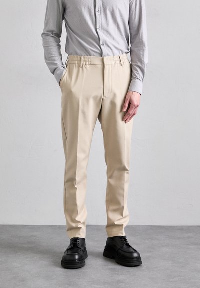 Beige tailored pants with a smooth finish, featuring an elastic waistband, side pockets, and straight legs, paired with black lace-up shoes.