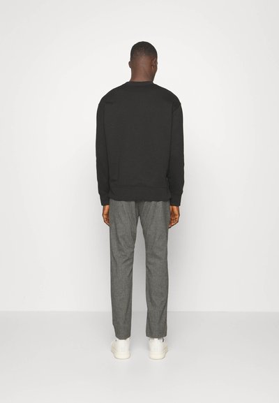 Schwarzer Sweatshirt mit rundem Halsausschnitt, kombiniert mit grauen gemusterten Hosen und weißen Sneakers, von hinten vor einem weißen Hintergrund betrachtet.