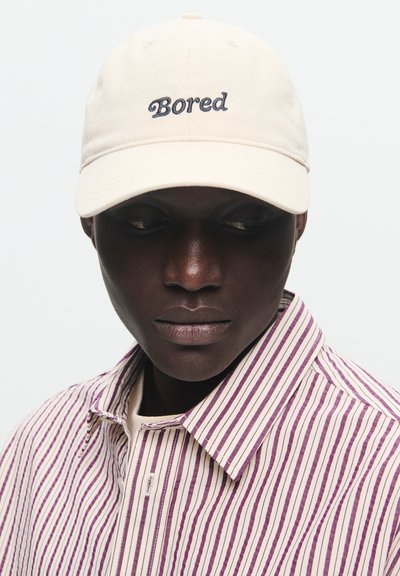 Šviesiai ruda kepurė su mėlynai siuvinėtu žodžiu "Bored". Dėvima su vertikaliai dryžuota marškinėliu su rožine ir kremine spalvomis.
