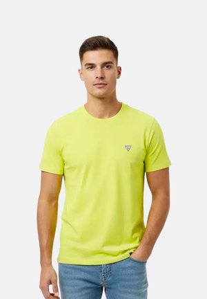 Homme portant un t-shirt jaune vif à manches courtes avec un petit logo sur la poitrine et un jean bleu, debout avec une main dans la poche.
