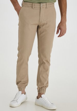 Bas du corps d'une personne portant un pantalon de jogging kaki avec des poignets élastiques et des baskets blanches, debout devant un fond uni.