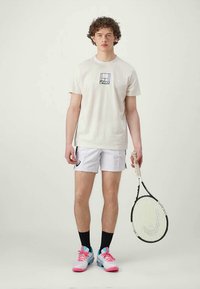 Camiseta de algodón blanca con un gráfico en azul y negro, combinada con pantalones cortos blancos. Lleva zapatillas de tenis rosas y azules, sosteniendo una raqueta de tenis.