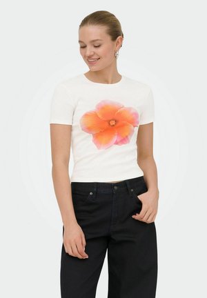 Mujer con cabello castaño claro recogido en un moño bajo, vestida con camiseta blanca con un gran estampado de flores naranja-rosa y pantalones negros, sonriendo con los ojos cerrados.