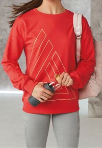 Rode sweatshirt met gouden geometrisch patroon, grijze leggings met strepen, houdt een grijze waterfles vast en heeft een lichtroze tas over de schouder.