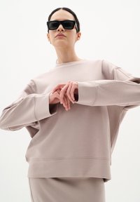 Ljust rosa sweatshirt i mjukt material, med sänkta axlar och vida ärmar. Modellen bär svarta fyrkantiga solglasögon.