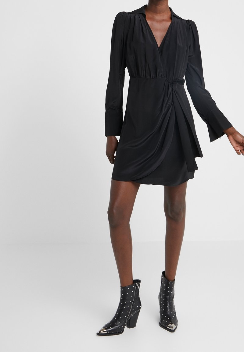 Robe portefeuille noire avec manches longues, encolure en V et taille ceinturée. Tissu lisse, détail noué sur le côté et jupe fluide.