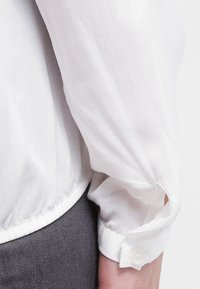 Camisa blanca de manga larga con dobladillo fruncido, que presenta un puño abotonado y una textura de tela suave y ligera. Vista en primer plano de la manga.