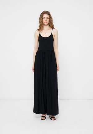 Γυναίκα με κατσαρά μαλλιά, φορώντας μαύρο μακρύ φόρεμα χωρίς μανίκια και μαύρα σανδάλια, που στέκεται μπροστά σε λευκό φόντο.