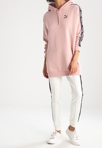 Rosa huvtröja med ficka fram, svart logotyp på bröstet och kontrasterande sidostreck. Matcher med vita joggers med svarta detaljer.