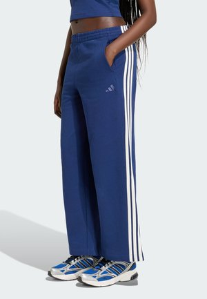 Osoba nosząca niebieskie spodnie dresowe Adidas z białymi paskami i niebieskie buty sportowe, stojąca z jedną ręką w kieszeni.