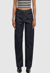 Frau trägt dunkelblaue Jeans mit geradem Bein, schwarze Knöchelstiefel und ein schwarz-weiß gestreiftes ärmelloses Cropped Top vor schlichtem Hintergrund.