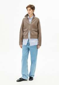 Bruine jas van een wolmix met een hoge kraag, rits aan de voorkant en twee grote zakken, gedragen over een blauw-wit gestreept shirt en spijkerbroek.