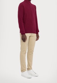 Burgunderfarbener Zip-Hals-Pullover aus Baumwollmischung; beige Slim-Fit-Hosen; weiße Sneaker. Der Pullover hat gerippte Bündchen und einen Saum; Logo auf der Brust.