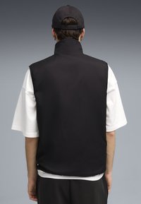 Schwarze ärmellose Weste mit hohem Kragen, aus glattem Stoff gefertigt. Über einem weißen T-Shirt getragen. Schlichtes Design, keine sichtbaren Muster oder Hardware-Details.