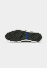 Semelle de chaussure noire avec un bord blanc et un motif triangulaire texturé, avec un petit logo rectangulaire bleu près du centre.