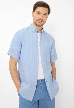 Mann trägt hellblaues kurzärmeliges Hemd über weißem T-Shirt, blaue Hose, steht mit den Händen in den Taschen vor weißem Hintergrund.