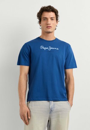 Junger Mann mit lockigem Haar, der ein blaues Pepe Jeans London T-Shirt und helle Jeans trägt, steht vor einem schlichten hellen Hintergrund.