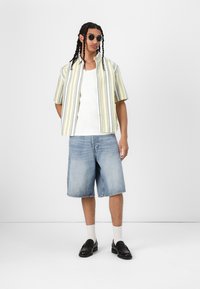 Camicia a maniche corte con bottoni, a righe gialle e grigie, abbinata a pantaloni corti in denim blu chiaro. Mocassini neri e calze bianche completano l'outfit.