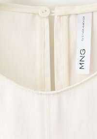 Gros plan sur l'encolure d'un vêtement blanc avec une petite fermeture par bouton et une étiquette indiquant MNG, taille EU 57 USA 4 MEX CH.
