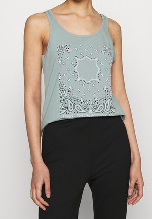 Žena nosící zelený krátký top bez rukávů se vzorem paisley na bílém a černém šátku, v kombinaci s černými kalhotami.