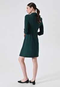 Dunkelgrünes Kleid mit einem Blazer-Kragen und Dreiviertelärmeln, das eine taillierte Form und einen nahtlosen Rücken aufweist, kombiniert mit schwarzen Ballerinas.