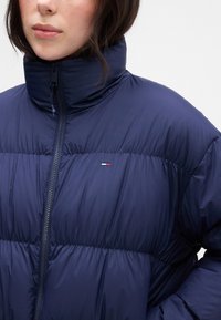 Granatowa kurtka puchowa z wysokim kołnierzem, zapinana na zamek, o pikowanej fakturze. Posiada małe, trójkolorowe logo na lewej piersi.