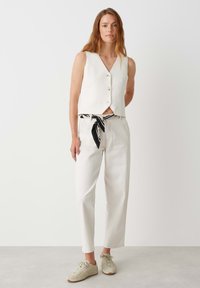 Débardeur blanc sans manches avec des boutons sur le devant, assorti à un pantalon blanc taille haute et une ceinture à nœud à motif noir et blanc, ainsi que des baskets légères.