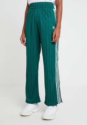 Person iført grønne Adidas træningsbukser med lodrette nålestriber og hvide sidelængder kombineret med hvide sneakers mod en ensfarvet baggrund.