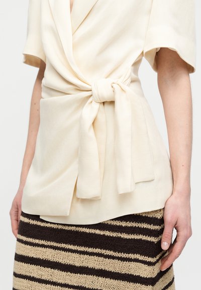 Επάνω μέρος τύπου wrap σε κρεμ απόχρωση με κοντά μανίκια και κόμπο στο μπροστινό μέρος, συνδυασμένο με πλεκτή ριγέ φούστα σε καφέ και μπεζ αποχρώσεις. Υφή από ύφασμα με χαλαρή εφαρμογή.