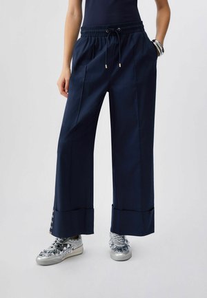 Osoba nosící tmavě modré kalhoty širokých nohavic s ohrnutými lemy a stříbrné tenisky s flitry, stojící před jednoduchým světlým pozadím.