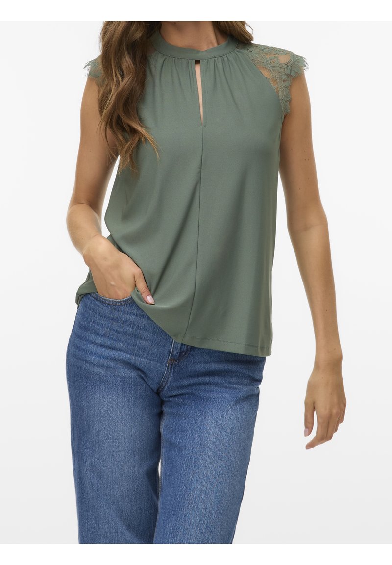 Femme portant un haut vert sauge sans manches avec des manches courtes en dentelle et une ouverture en forme de trou de serrure à l'avant, associé à un jean bleu, une main dans la poche.