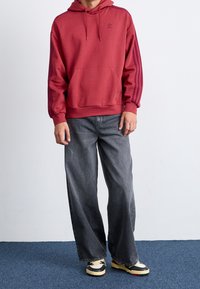 Röd hoodie med en framficka och dragsko på huvan, med tre vertikala ränder på ärmarna. Kombinerad med lösa grå jeans och sneakers.