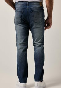 Vaqueros de mezclilla azul oscuro con corte ajustado, cinco bolsillos y un ligero desgastado. La cintura cuenta con una etiqueta de cuero. Se llevan zapatillas blancas.