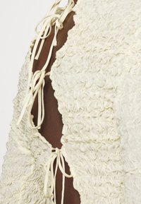 Detailní záběr na hnědou pleť viditelnou skrze otevřená záda krémového topu texturovaného látkou, který je zajištěn několika tenkými šňůrkami.