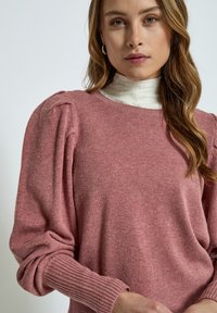 Pink knitted sweater with puffed shoulders, ribbed cuffs, and a soft texture. Worn over a white high-neck top.