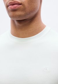 Λευκό βαμβακερό T-shirt με στρογγυλή λαιμόκοψη· διαθέτει μικρό, ανάγλυφο λογότυπο «CK» στο αριστερό στήθος· λεία υφή, μινιμαλιστικός σχεδιασμός.