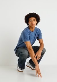 Sinine ümar kaelusega t-särk, lühikesed varrukad, kerge kangas. Tumedad teksad, hõbedased tossud. Mudel põlvitab, lokkis juustega, ühevärvilise taustaga.