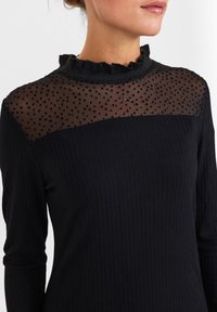 Schwarzes, geripptes Langarmoberteil mit einem durchsichtigen, gepunkteten Netz-Einsatz und einem Rüschenausschnitt. Glatte Textur, taillierte Form und fester Oberteil.