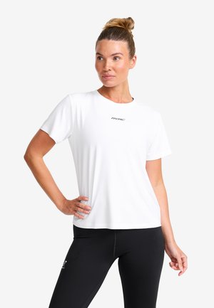 Frau mit einem Dutt, die ein weißes Kurzarmhemd mit dem Text "RVRC" und schwarze Leggings trägt, steht mit einer Hand auf der Hüfte.
