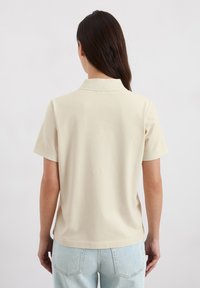 Beige katoenen poloshirt met een kraag, korte mouwen en een ontspannen pasvorm, gecombineerd met lichtblauwe jeans.