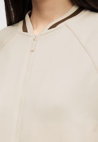 Femme portant une veste beige zippée avec un col côtelé présentant des rayures marron et crème, vue du cou jusqu'au milieu de la poitrine.