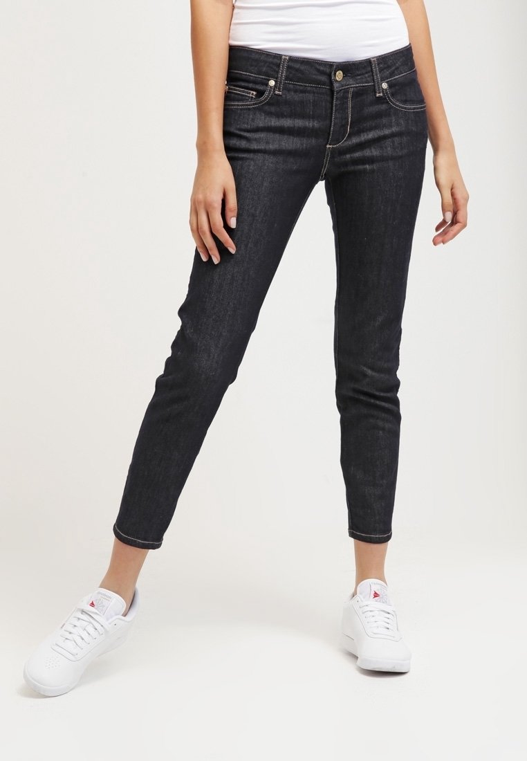 Jeans ajustados de color azul oscuro, hechos de denim, con cinco bolsillos, costuras doradas y un ligero desvanecimiento. Combinados con zapatillas blancas.