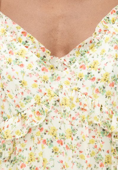 Κοντινό πλάνο στo ντεκολτέ μιας φλοράλ φόρεμα με βολάν σε κίτρινο, ροζ και πράσινο σε λευκό ύφασμα, που φοριέται από άτομο με μέτριο τόνο δέρματος.