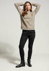 Frau trägt beigen Pullover, schwarze Skinny Jeans und schwarze Stiefeletten, steht mit Händen hinter dem Kopf vor einem einfarbigen Hintergrund.