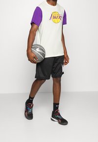 Camiseta de los Los Angeles Lakers con mangas moradas, pantalones cortos negros y zapatillas de Nike multicolores; sosteniendo un balón de baloncesto gris con el logo de Nike.