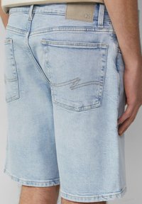 Shorts en denim bleu clair avec une coupe décontractée, comportant deux poches arrière et un détail de couture en zigzag subtil. Étiquette de taille en cuir beige.
