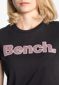Camiseta negra con cuello redondo, que presenta la palabra "Bench." en letras gruesas de color blanco y burdeos con puntos. Textura de algodón suave.